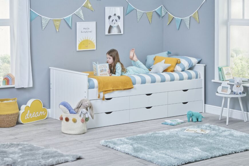 girl is laying and reading a book on the wooden white bed that has storage space in kids room